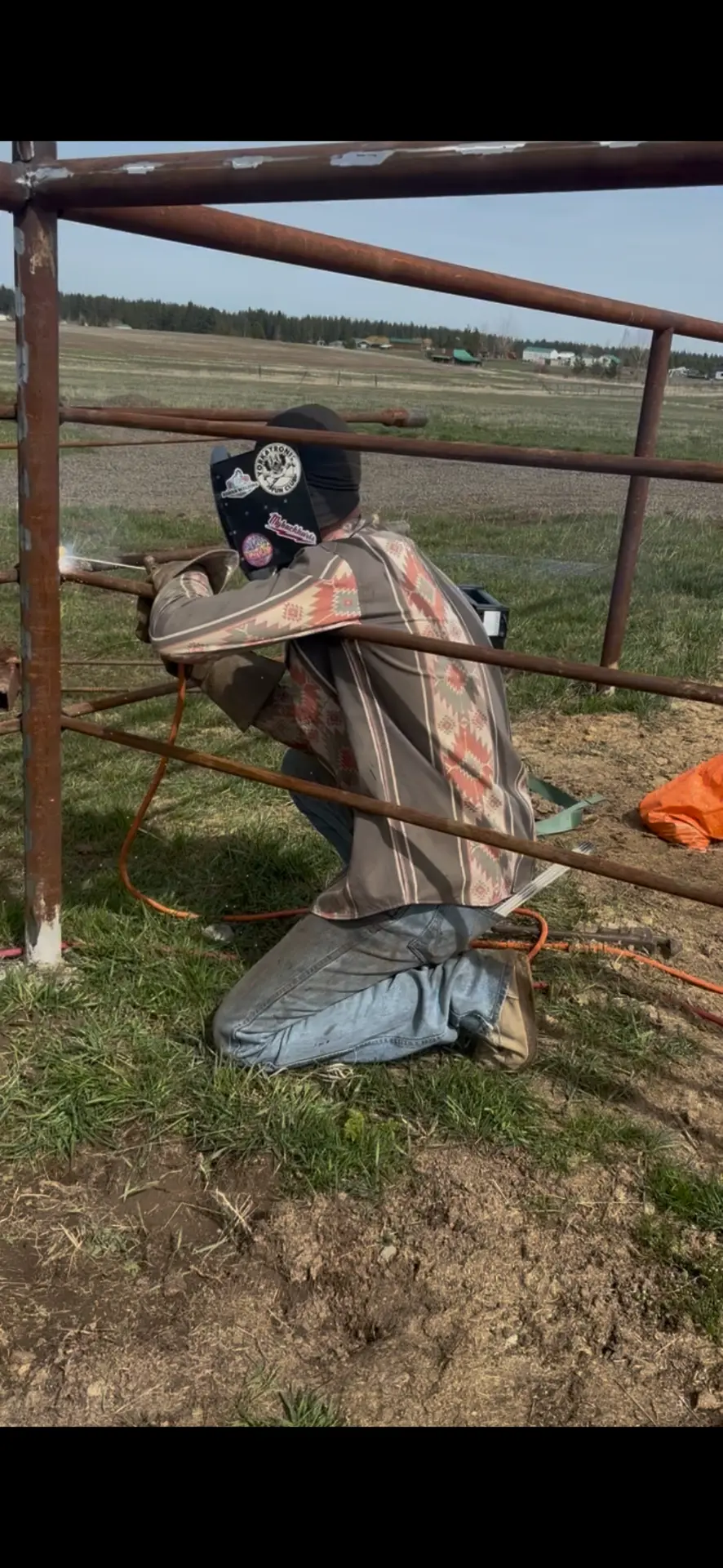 Welder kneeling in a ranch pasture welding steel pipe corral rails